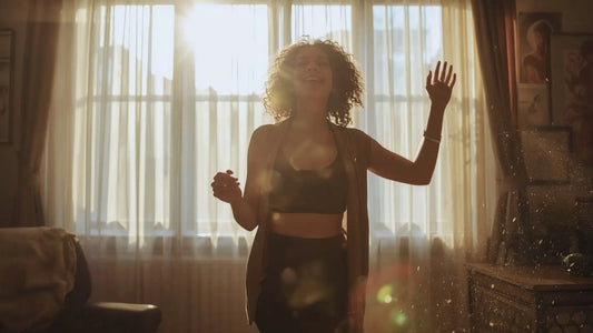 Une femme souriante et pleine d'énergie qui danse dans son salon ou la lumière du soleil entre par des rideaux fermés